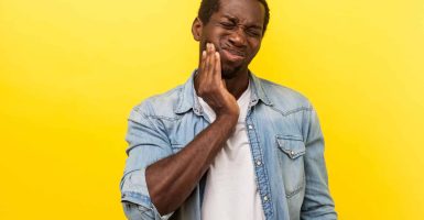 Terrible toothache. Portrait of unhealthy man in denim casual shirt rubbing his cheek and grimacing from acute pain, teeth problems, dental injury. indoor studio shot isolated on yellow background