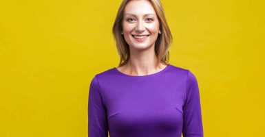 Portrait of positive elegant woman in tight purple dress standing, looking at camera with cute engaging toothy smile, beautiful with clean shiny skin. indoor studio shot isolated on yellow background