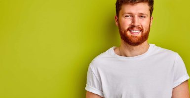 young man in casual t-shirt smiling at camera, handsome guy with red hair and beard isolated over green background