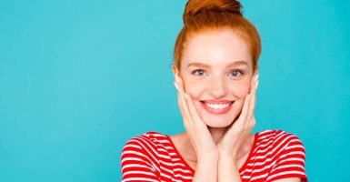 Close-up portrait of nice cute attractive adorable magnificent lovely cheerful optimistic red-haired girl with bun, beaming smile, holding cheeks in palms, isolated on bright vivid blue background