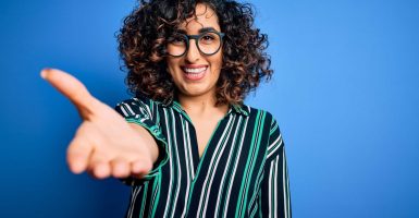 Young beautiful curly arab woman wearing striped shirt and glasses over blue background smiling friendly offering handshake as greeting and welcoming. Successful business.