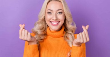 Photo of optimistic gorgeous girl with stylish hairdo dressed orange sweater fingers showing k-heart isolated on purple color background.
