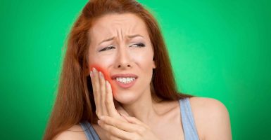 Closeup portrait young woman with sensitive toothache crown problem about to cry from pain touching red area outside mouth with hand isolated green background. Negative emotion face expression feeling
